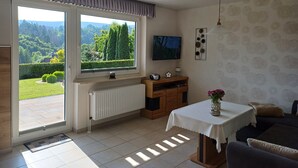 A living room with a large window, a television, a radiator, a sofa, and a small table with a vase.