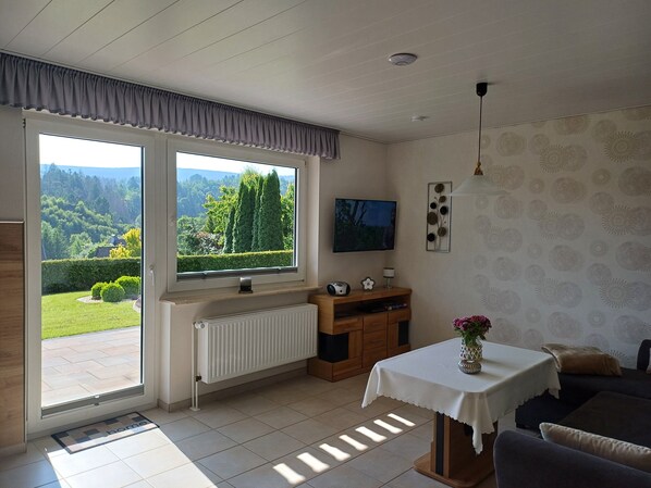 Un salon avec une grande fenêtre, un téléviseur, un radiateur, un canapé et une petite table sur laquelle est posé un vase.