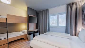 Una habitación de hotel moderna con una cama litera, una cama plana, un televisor montado en un panel de madera, una mesita y una ventana con cortinas.