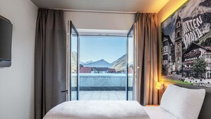 A hotel room with a bed, a large window, and a view of mountains and buildings. - B&B Hotel Mittenwald (Mittenwald)