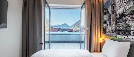 A hotel room with a bed, a large window, and a view of mountains and buildings.