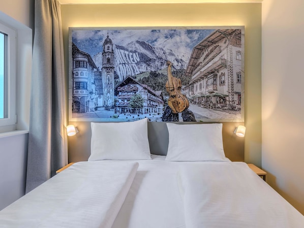 Habitación de hotel con cama, dos lámparas de noche y un mural grande que muestra un pueblo nevado en las montañas.