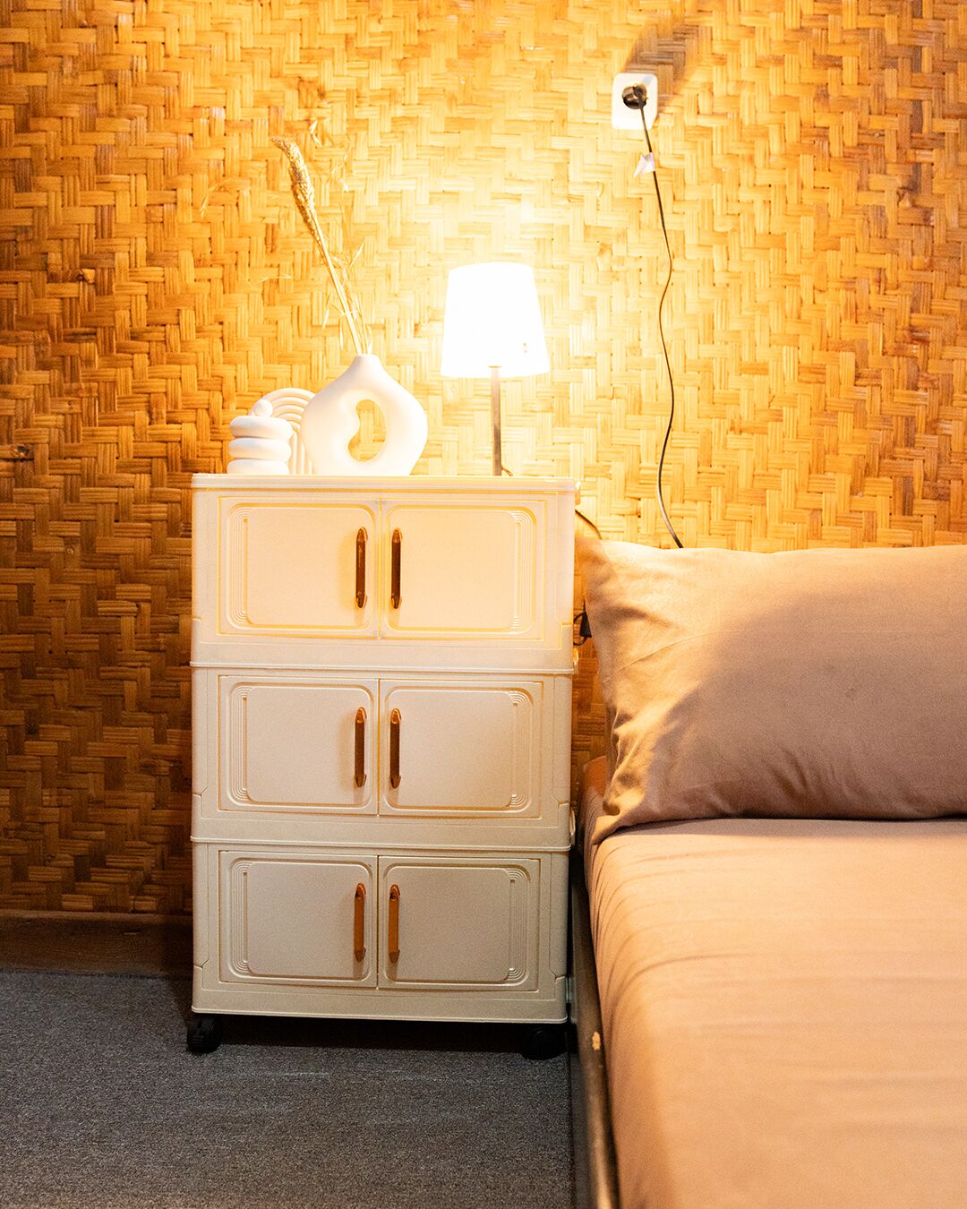 A white bedside table with a lamp and a vase, a beige bed, and a woven wall.