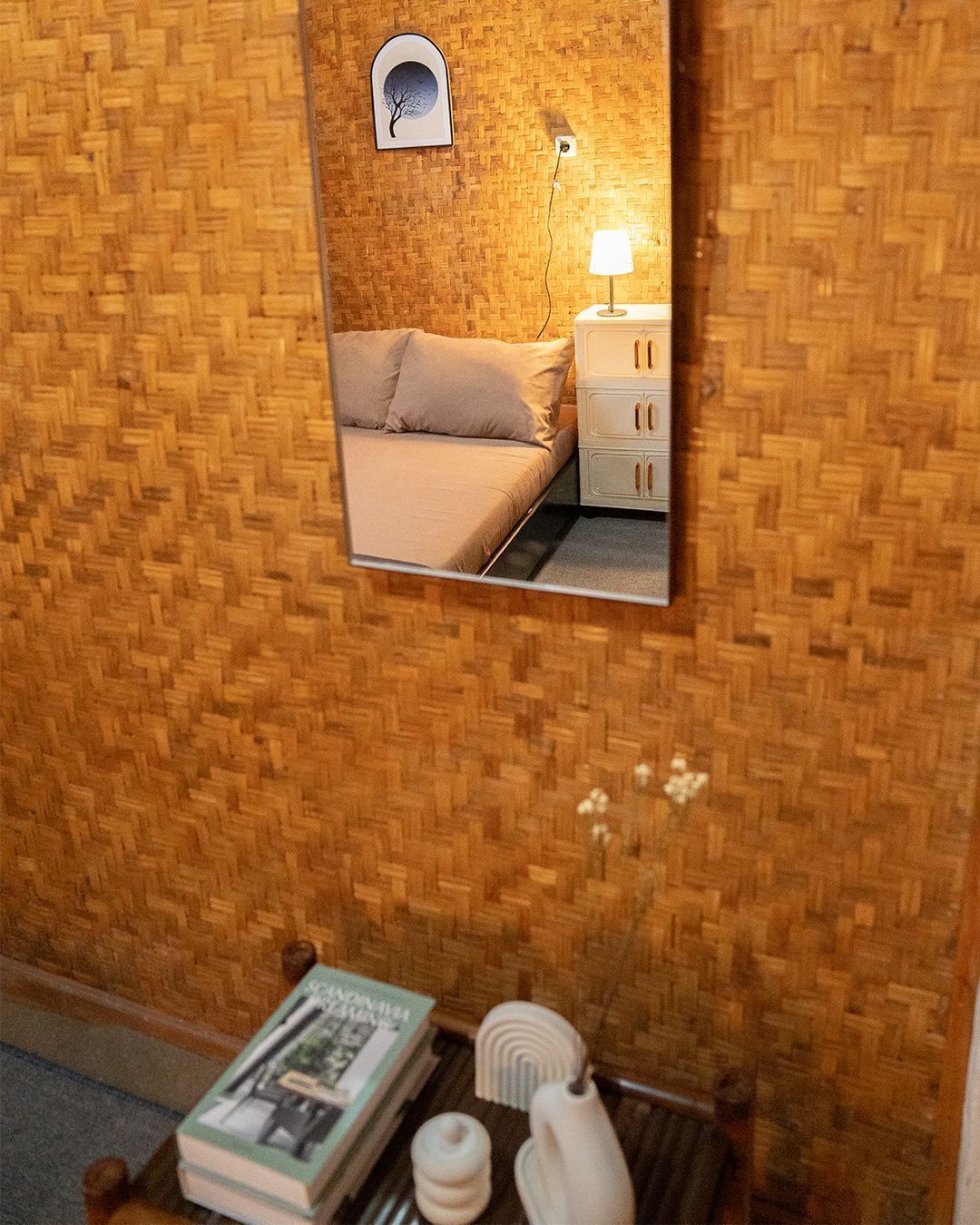 A room with a woven wall, a bed with beige bedding, a bedside table with a lamp, and a mirror reflecting the room.
