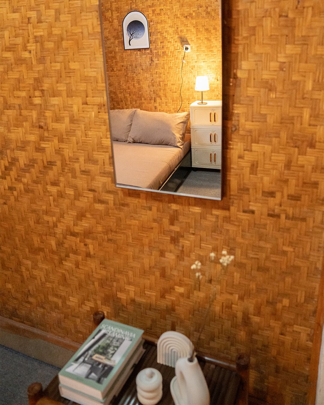 A room with a woven wall, a bed with beige bedding, a bedside table with a lamp, and a mirror reflecting the room.