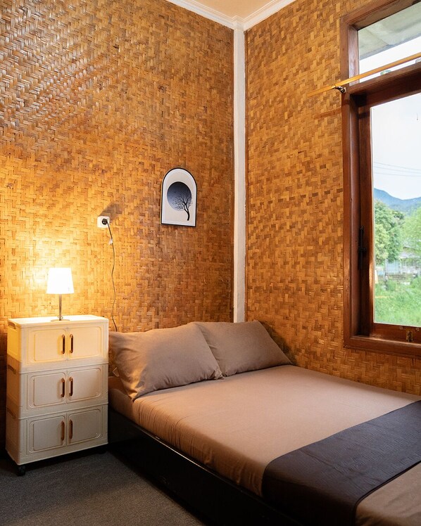 Une chambre avec un mur décoré d’un motif en chevrons en bois, un lit avec un matelas et des oreillers marron, une table de chevet avec une lampe, et une fenêtre donnant sur un paysage verdoyant.