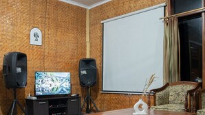 A room with a wooden wall, a large flat-screen TV, two speakers, a projector screen, a wooden table, and two patterned armchairs. - Villa Situ Patenggang BKSDA (Rancabali)