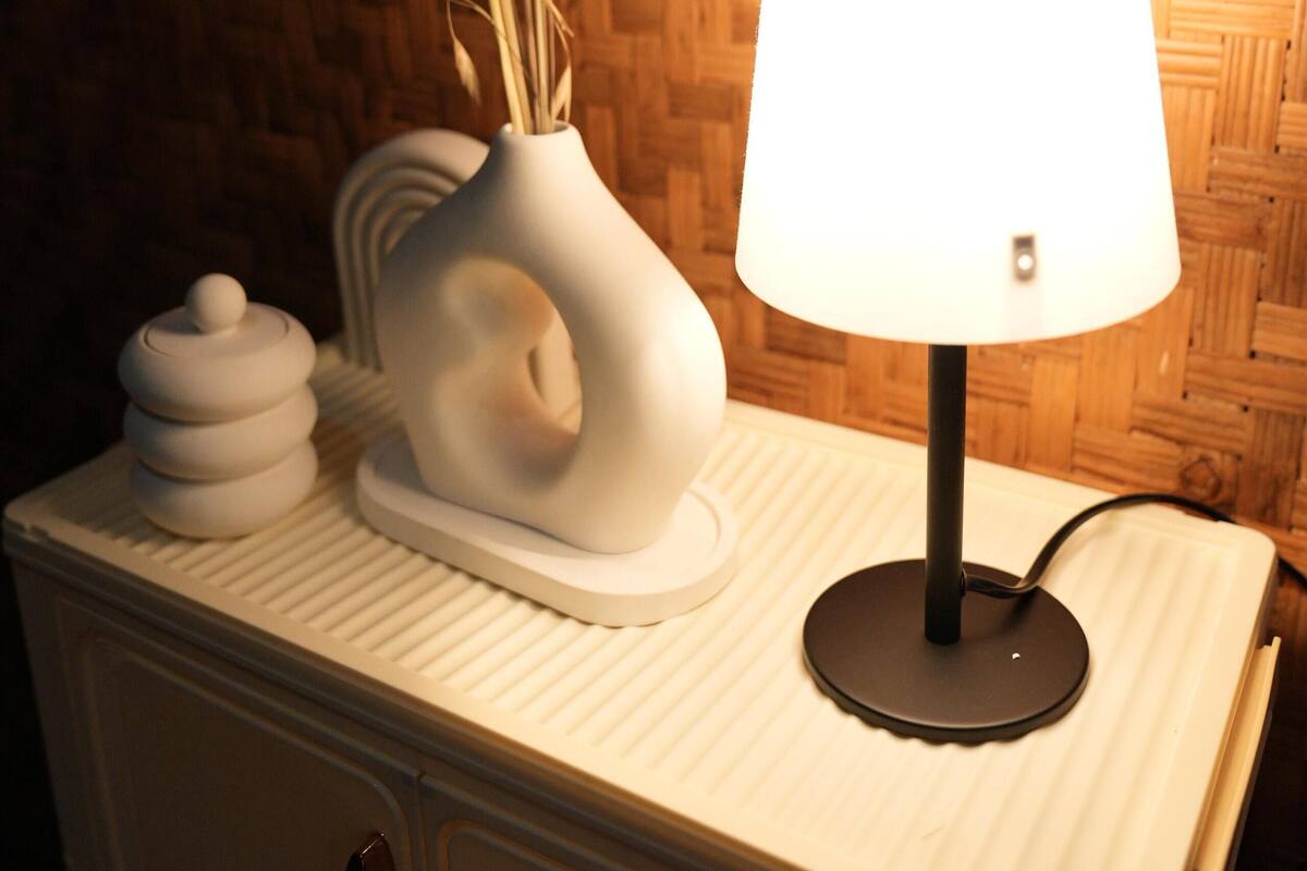 Une table de chevet avec une lampe, un vase contenant des plantes séchées et une pile de récipients blancs.