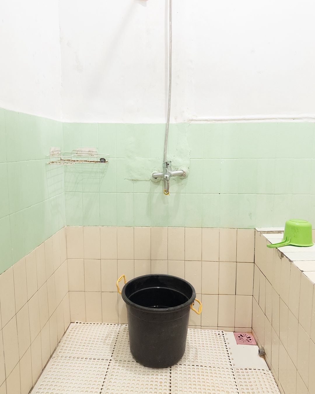 A small, tiled bathroom with a shower, a black bucket, and a green container on the counter.