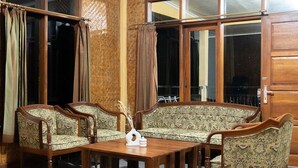 A corner room with a wooden floor, a sofa set, a coffee table, and large windows with curtains. - Villa Situ Patenggang BKSDA (Rancabali)