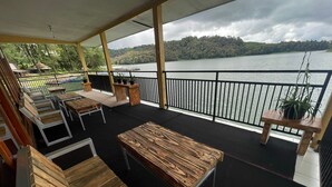 A covered outdoor seating area with wooden furniture, a railing, and a view of a lake and forested hills. - Villa Situ Patenggang BKSDA (Rancabali)