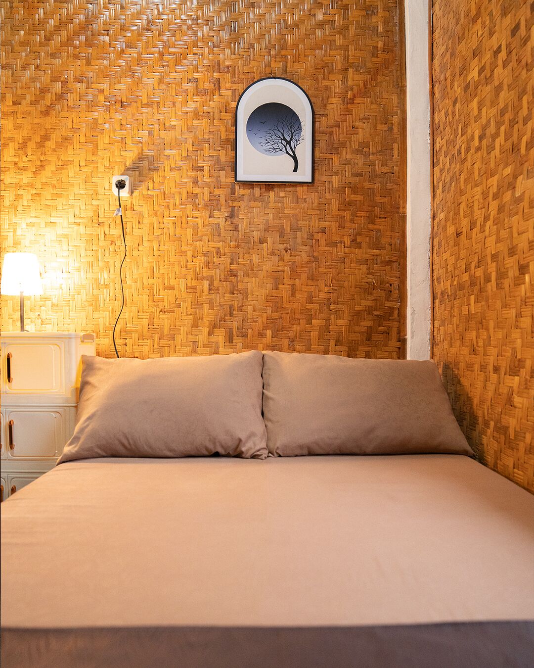 A room with a woven wall pattern, a bed with beige bedding, a nightstand with a lamp, and a framed picture of a tree.