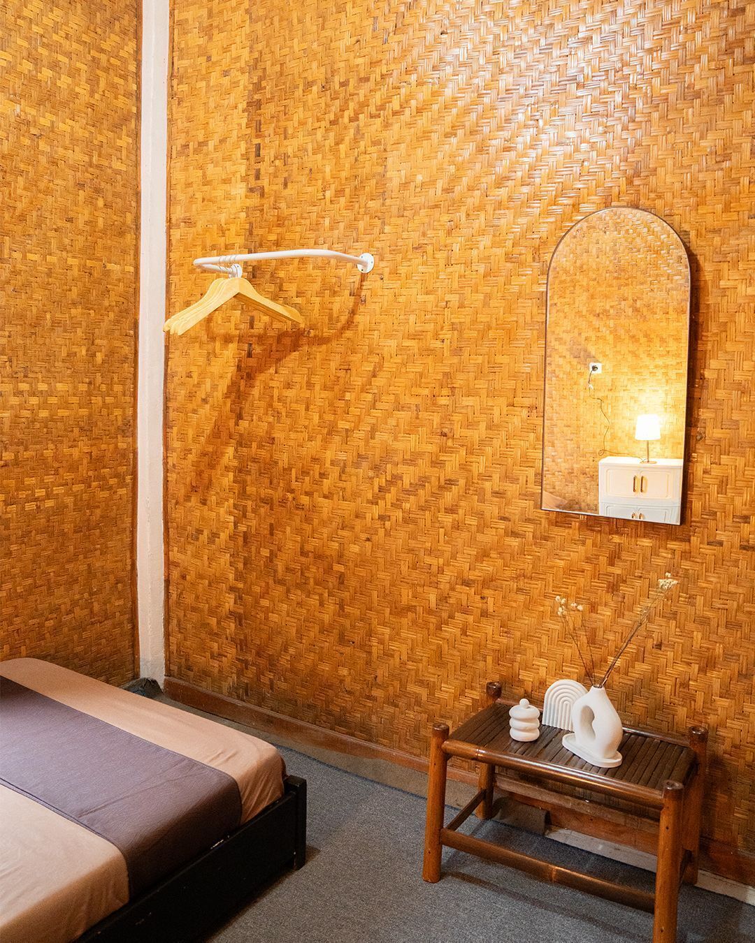 A room with a bed, a wooden wall, a mirror, a small table with a lamp, and a towel rack.