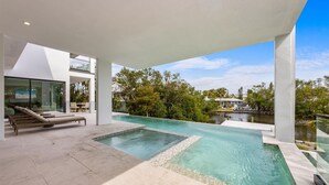 Un área de piscina moderna al aire libre con sillones de descanso, una sala con paredes de vidrio y vistas a la vegetación y al agua circundantes.