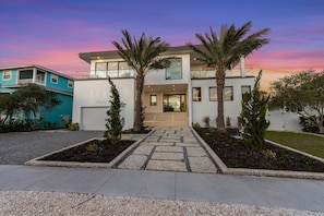 A modern two-story house with a stone pathway leading to the entrance, surrounded by palm trees and a well-maintained garden.