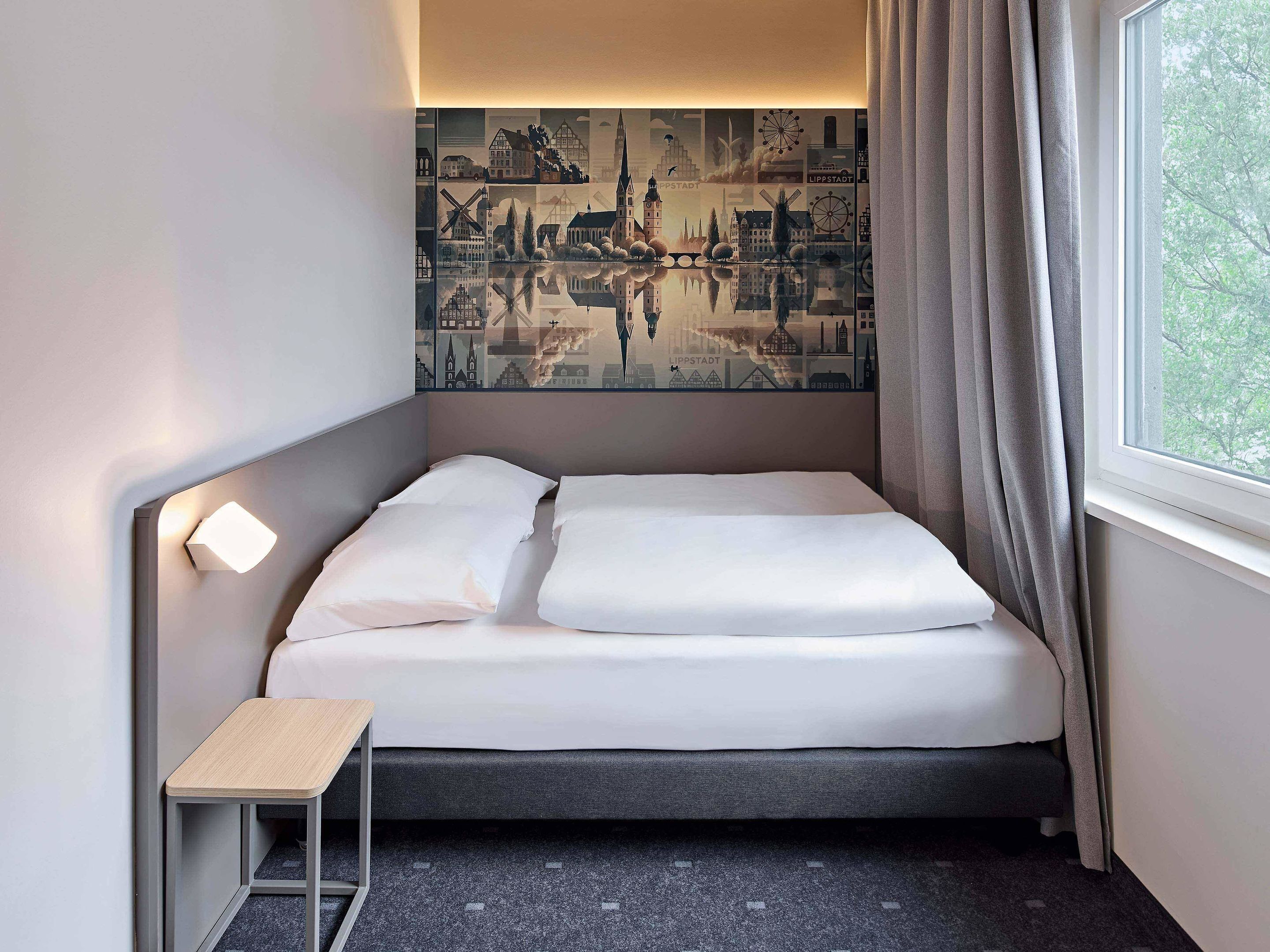 Een hotelkamer met een bed, een tafeltje, een wandlamp en een raam met gordijnen.