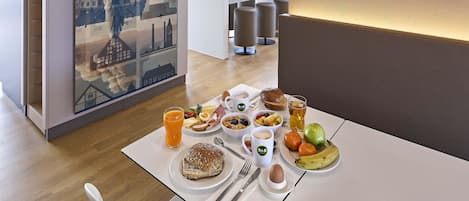 A breakfast setting with a table set for two, featuring a variety of foods and drinks, a cityscape mural, and modern lighting.