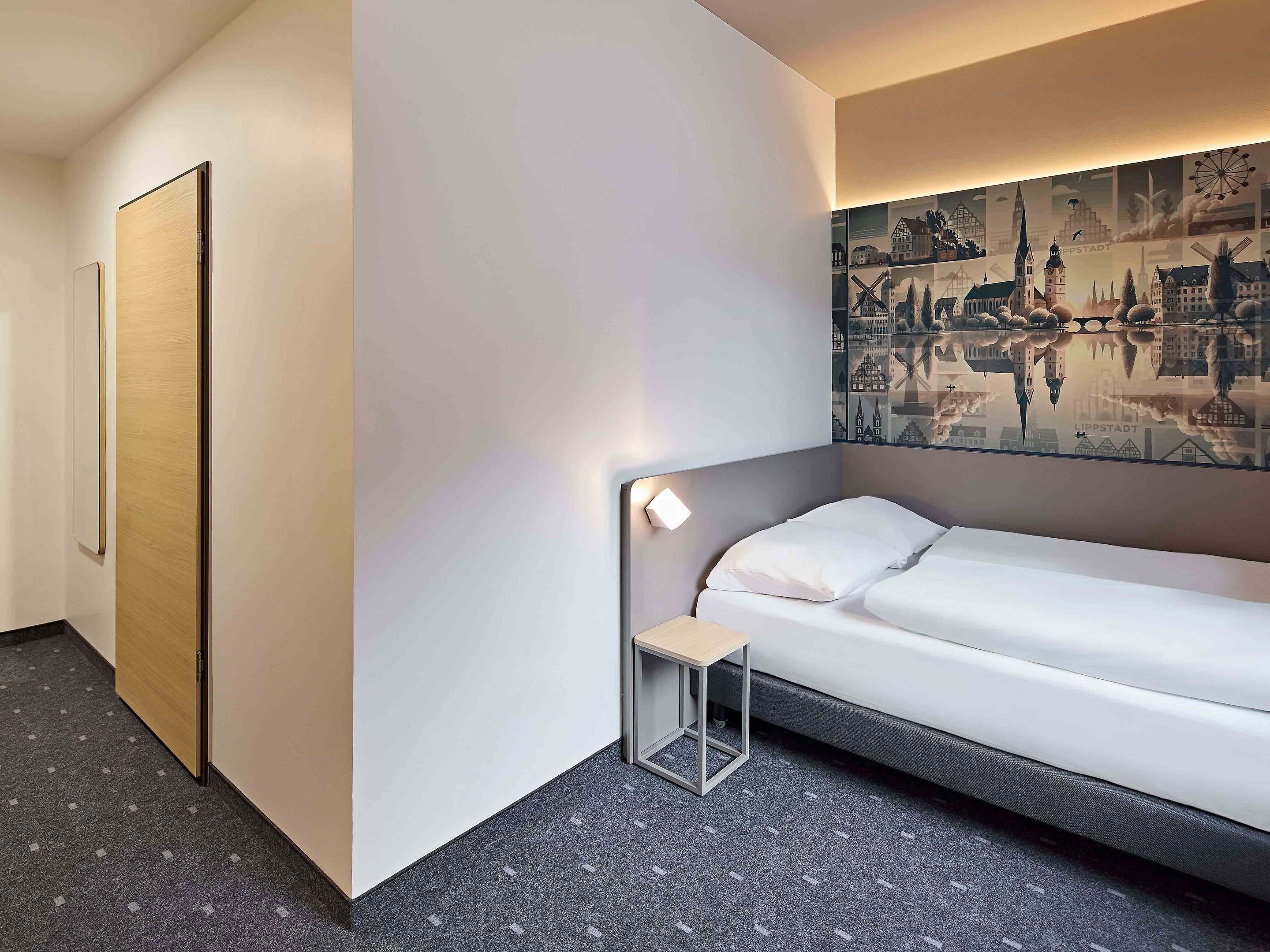 A hotel room with a single bed, a small bedside table, a wall-mounted light, and a cityscape mural.