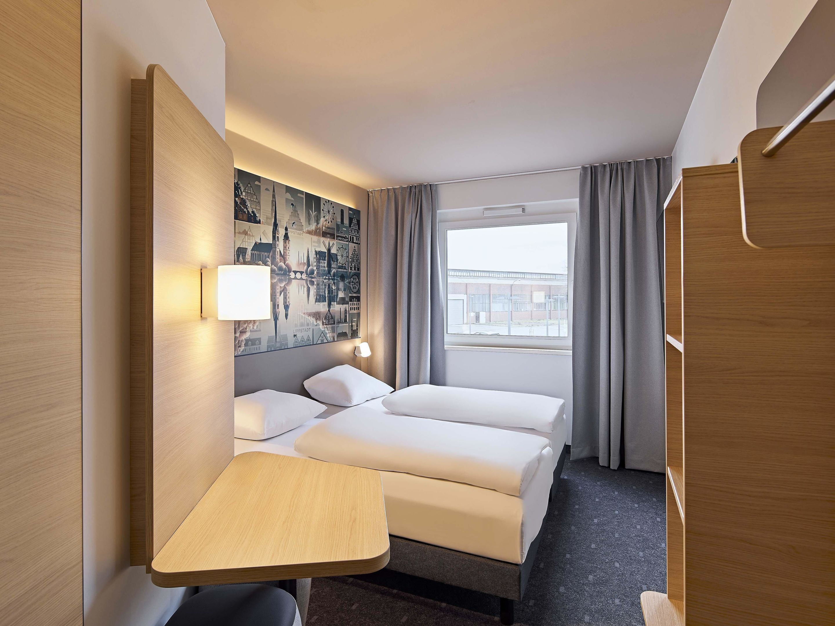 A hotel room with a bed, a desk, a lamp, and a large window with curtains.