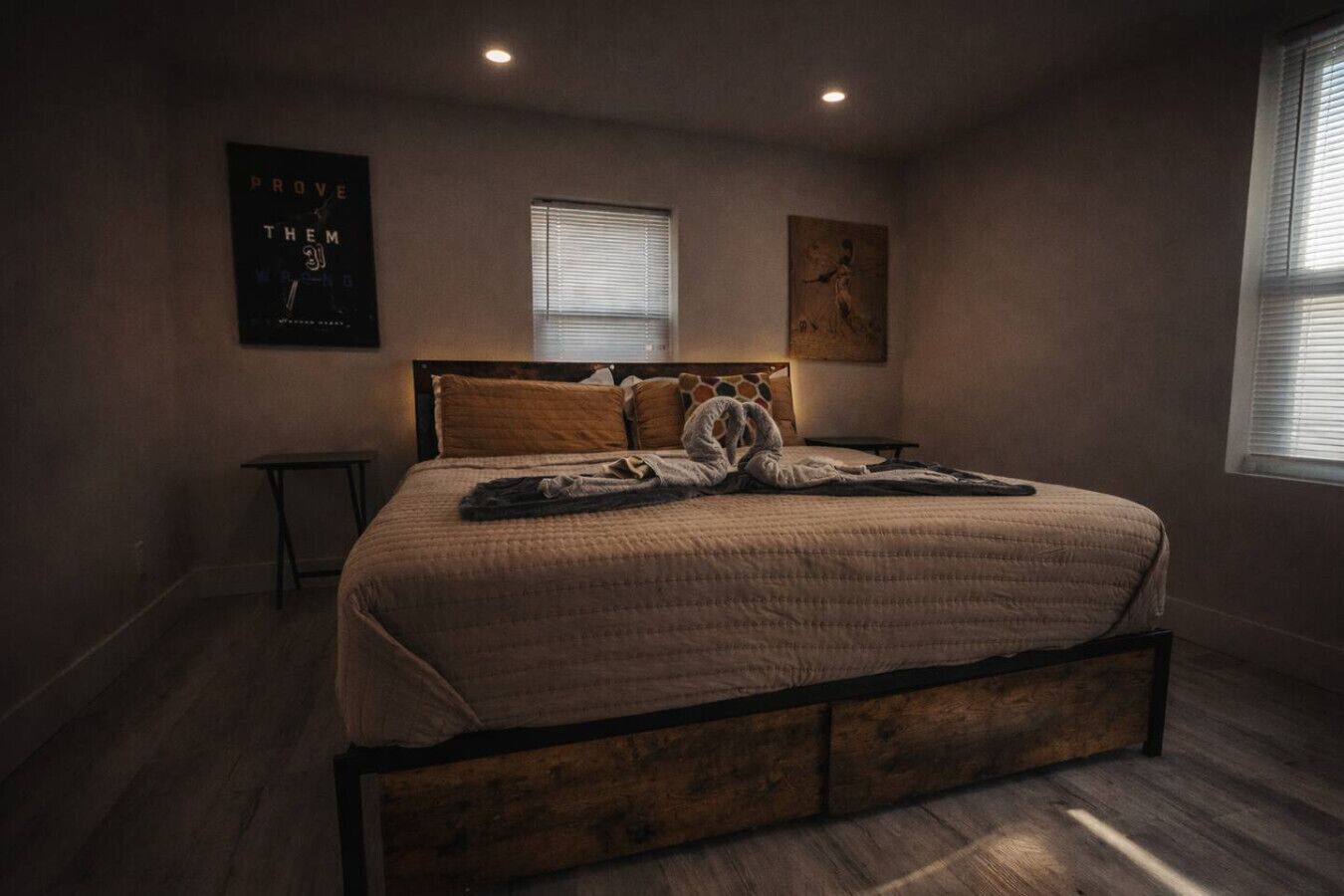 Ein Schlafzimmer mit einem hölzernen Bettgestell, einer großen Bettdecke, zwei Fenstern mit Jalousien, einem kleinen Tisch und Bildern an den Wänden.