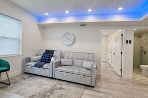 A modern living room with a grey sofa, a clock on the wall, and a glass shower area.