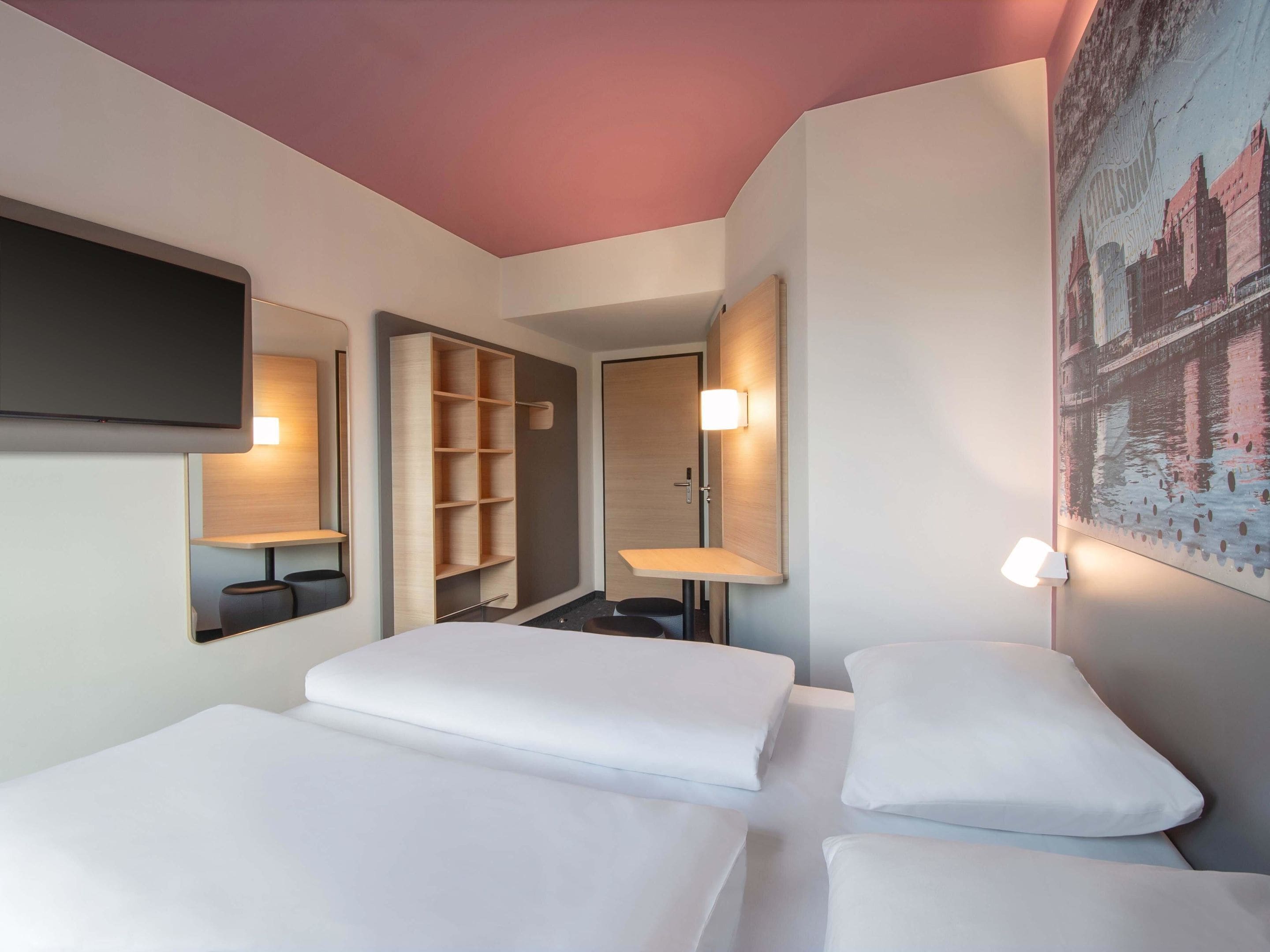 Une chambre d’hôtel avec deux lits, une télévision, un miroir, une étagère et un tableau.