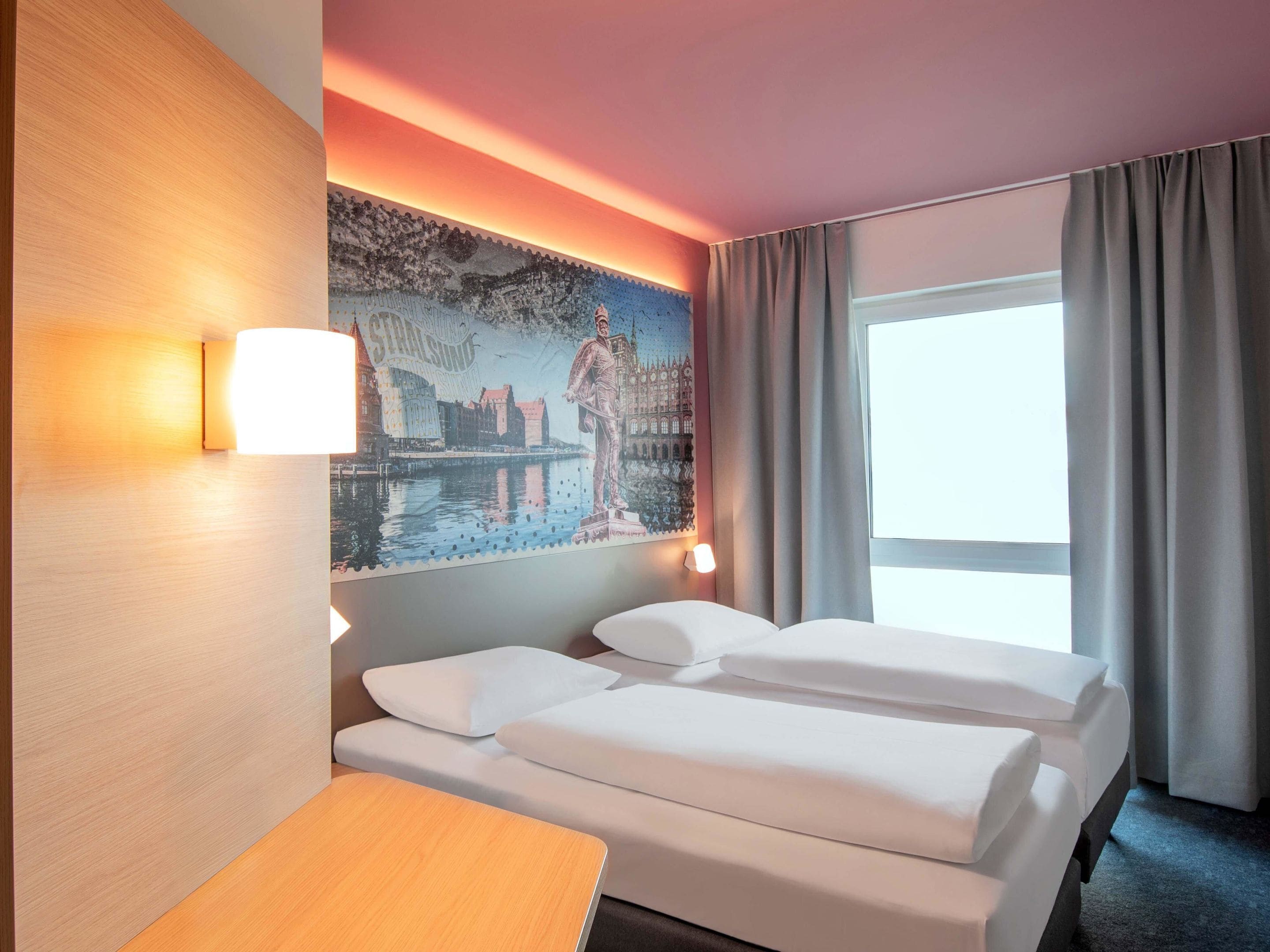 A hotel room with a bed, a desk, a lamp, a painting, and a window with curtains.