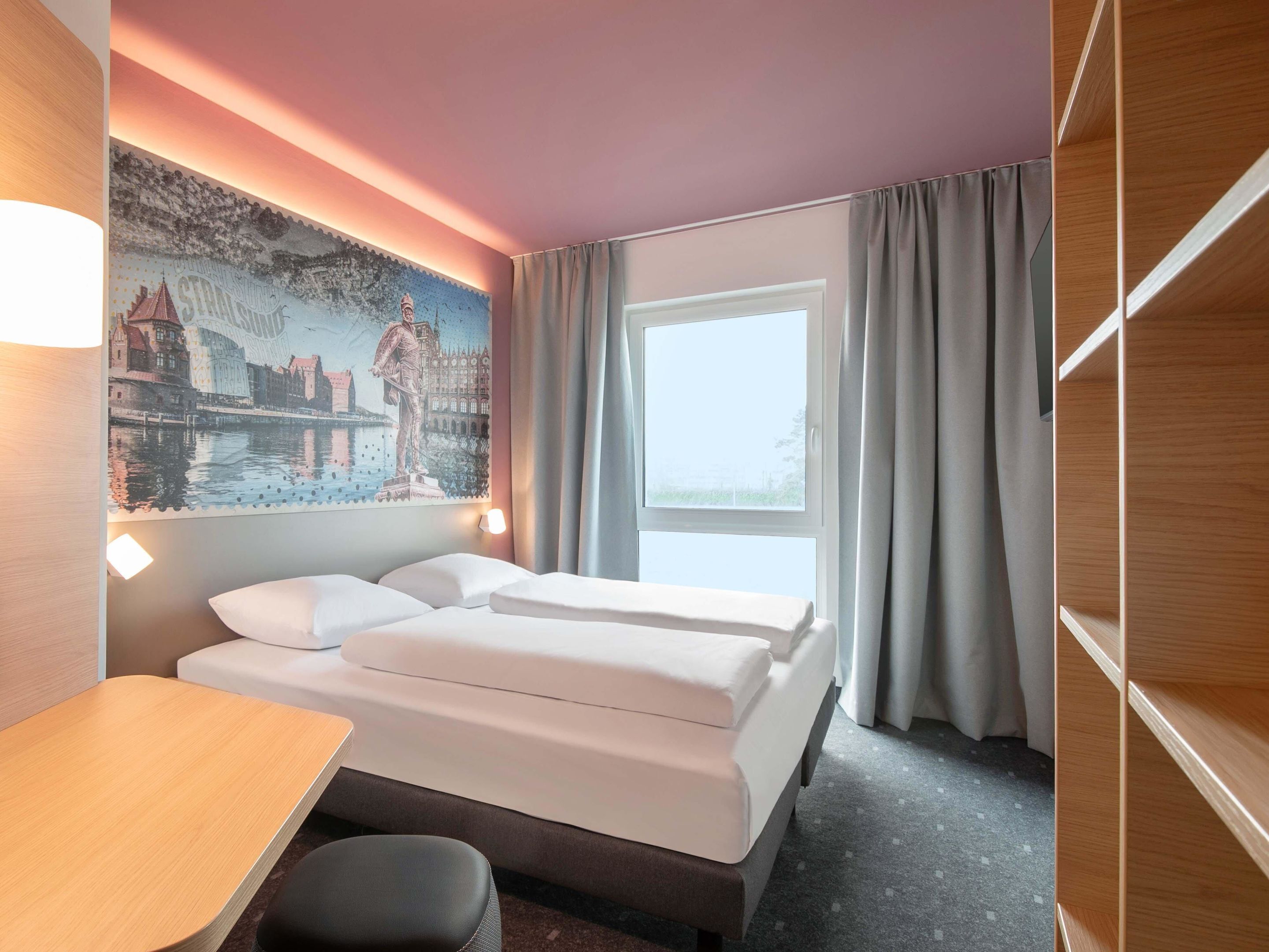 A hotel room with a bed, a desk, a chair, a large window, and a wall mural.