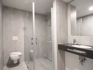 Egy modern fürőszoba üvegfalas zuhanyzóval, WC-vel és egy mosdókagylóval, mely felett tükör található.
