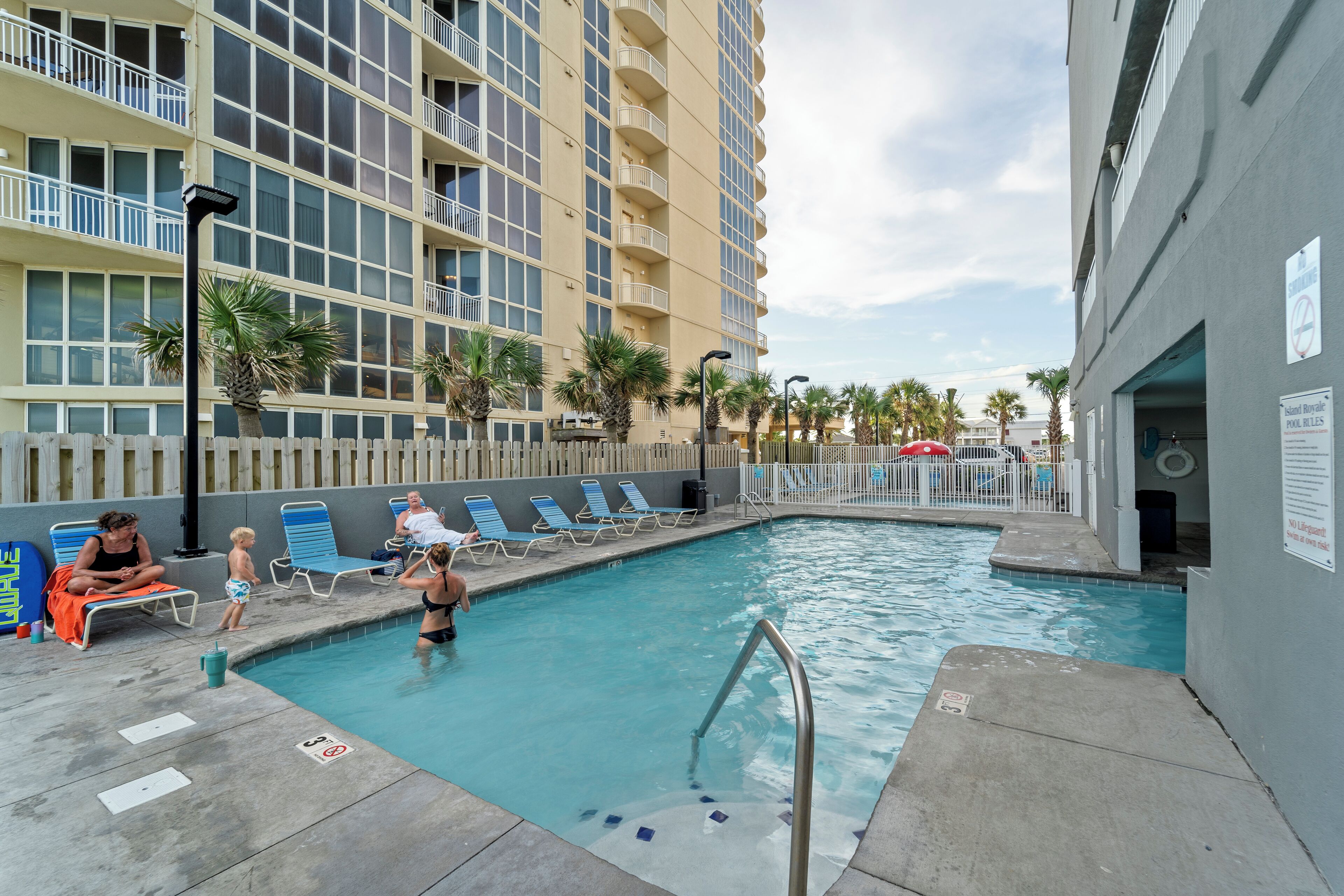 A swimming pool area with lounge chairs, a pool ladder, and a building with balconies in the background.