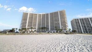 Un hotel sulla spiaggia con diversi piani, una spiaggia sabbiosa in primo piano e palme.