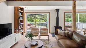 Ein Wohnzimmer mit Fernseher, Bücherregal, zwei Sesseln, einer Couch, einem Kamin und großen Fenstern.