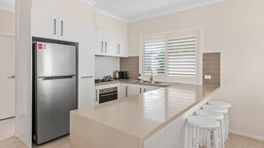 Cozinha moderna com geladeira de aço inoxidável, armários brancos, uma ilha grande com banquetas e uma janela com persianas.