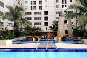 A swimming pool with a water slide, palm trees, and a building with multiple windows and balconies. - Studio and 1BR Bassura City City View (Min Stay 3 Nights) (East Jakarta)