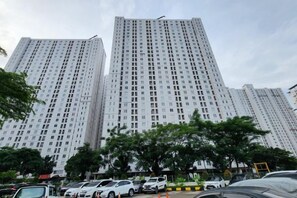 A high-rise building with multiple floors, surrounded by parked cars and trees. - Studio and 1BR Bassura City City View (Min Stay 3 Nights) (East Jakarta)