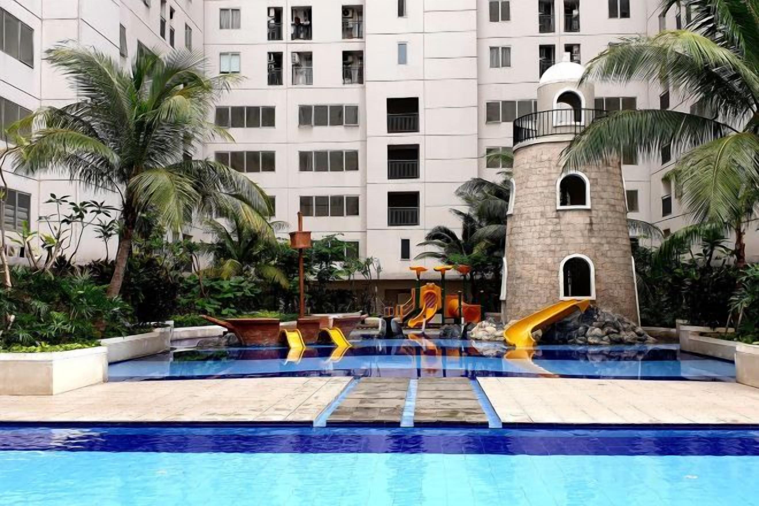 Uma piscina com escorregador, palmeiras e um edifício com várias varandas ao fundo.