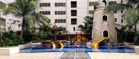 Uma piscina com escorregador, palmeiras e um edifício com várias varandas ao fundo.