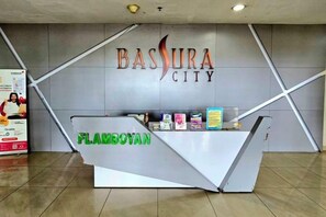 Ein Empfangsschalter mit einem Schild mit den Aufschriften 'BASURA CITY' und 'FLAMBOYAN' vor einer modernen Wand mit geometrischen Mustern.