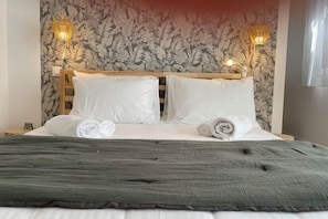 Un lit bien fait, avec du linge de lit blanc et deux serviettes enroulées, une couverture anthracite et une tête de lit en bois.
