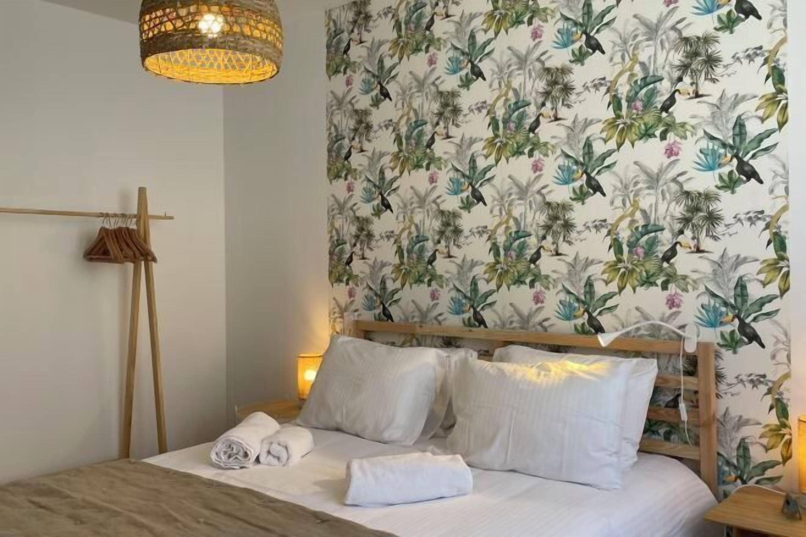 Una cama bien tendida con sábanas y almohadas blancas, un cabecero de madera y un papel pintado con motivos tropicales.