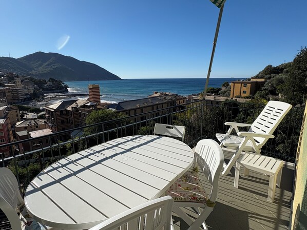 Un balcone con tavolo e sedie affaccia su una vista costiera con edifici e una montagna all'orizzonte.