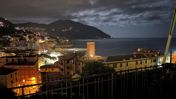 Une vue nocturne d’une ville côtière, avec des bâtiments illuminés, une tour à rayures bien visible et une vue sur la mer.