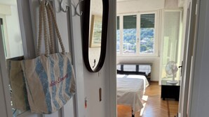 Une chambre avec un lit, une table de chevet sur laquelle se trouve un ventilateur, une fenêtre donnant sur un paysage agréable et un sac accroché au mur.