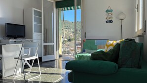 Une pièce avec un téléviseur posé sur un bureau, une chaise, un canapé donnant sur l’extérieur et une porte-fenêtre donnant sur un balcon.
