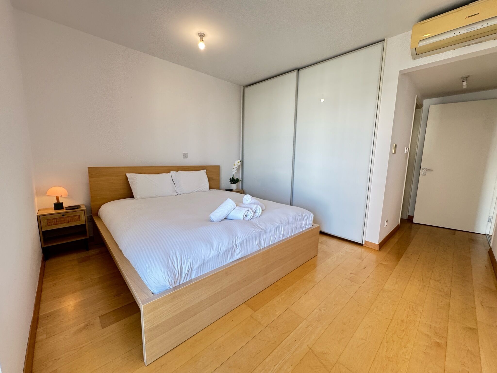 Um quarto com cama de madeira, mesa de cabeceira com luminária, porta de armário deslizante e unidade de ar condicionado instalada no teto.