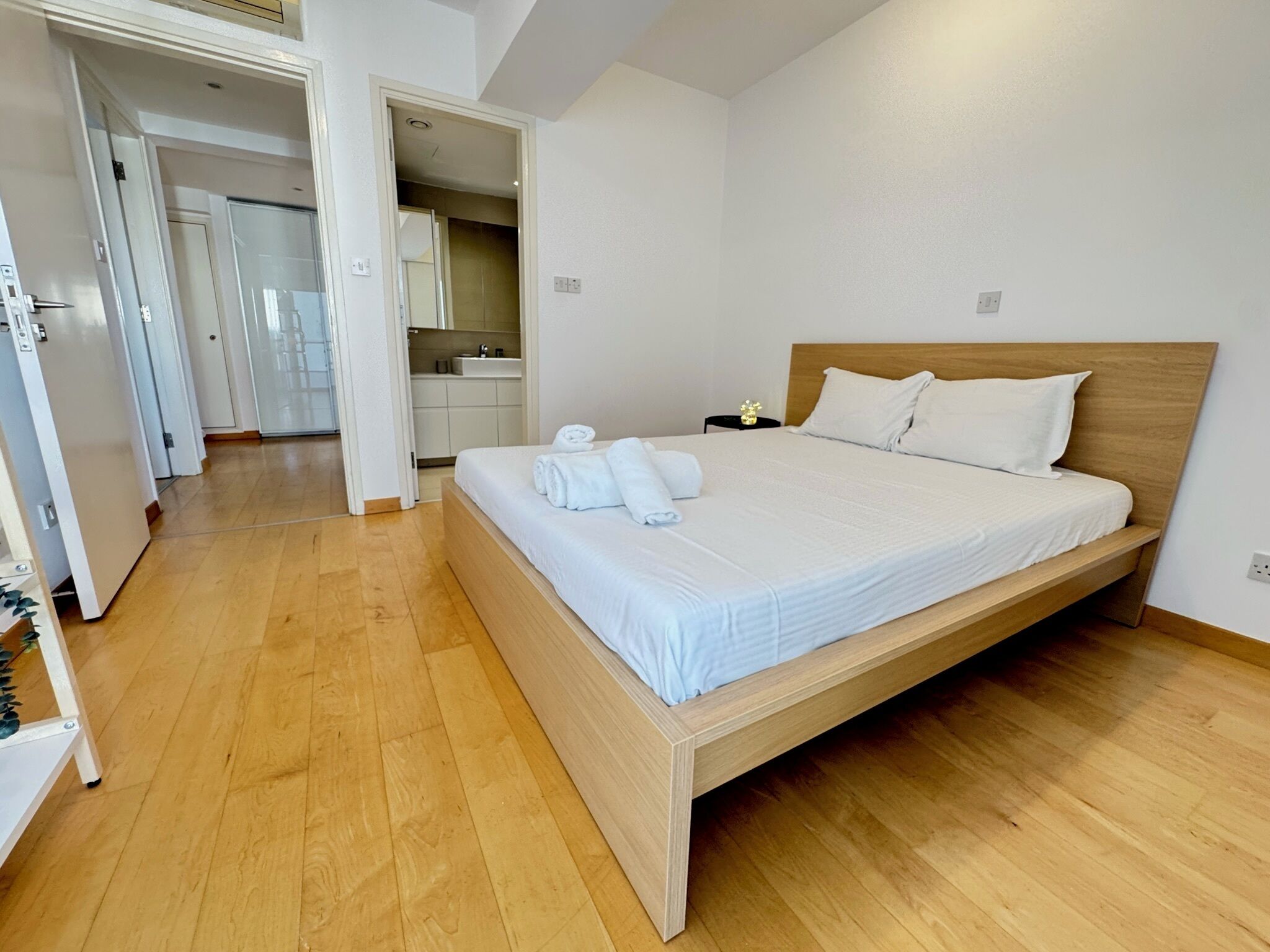 Habitación de hotel moderna con cama de madera, ropa de cama blanca y baño a la vista con lavamanos y espejo.