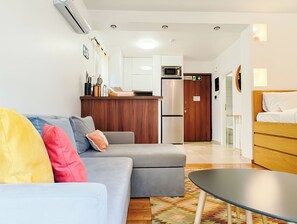 Un salon moderne avec un canapé gris, un meuble en bois, un petit coin cuisine équipé d’un micro-ondes et d’un réfrigérateur, et un lit avec une tête de lit en bois.