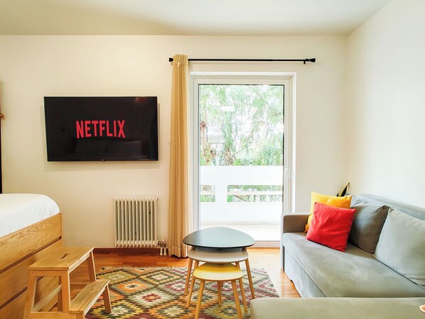 A room with a bed, a sofa, a small table, a radiator, a TV displaying 'NETFLIX', and a window with a view of greenery.