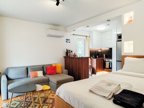A modern hotel room with a sofa, a small table, a bed, a kitchenette, and an air conditioning unit.