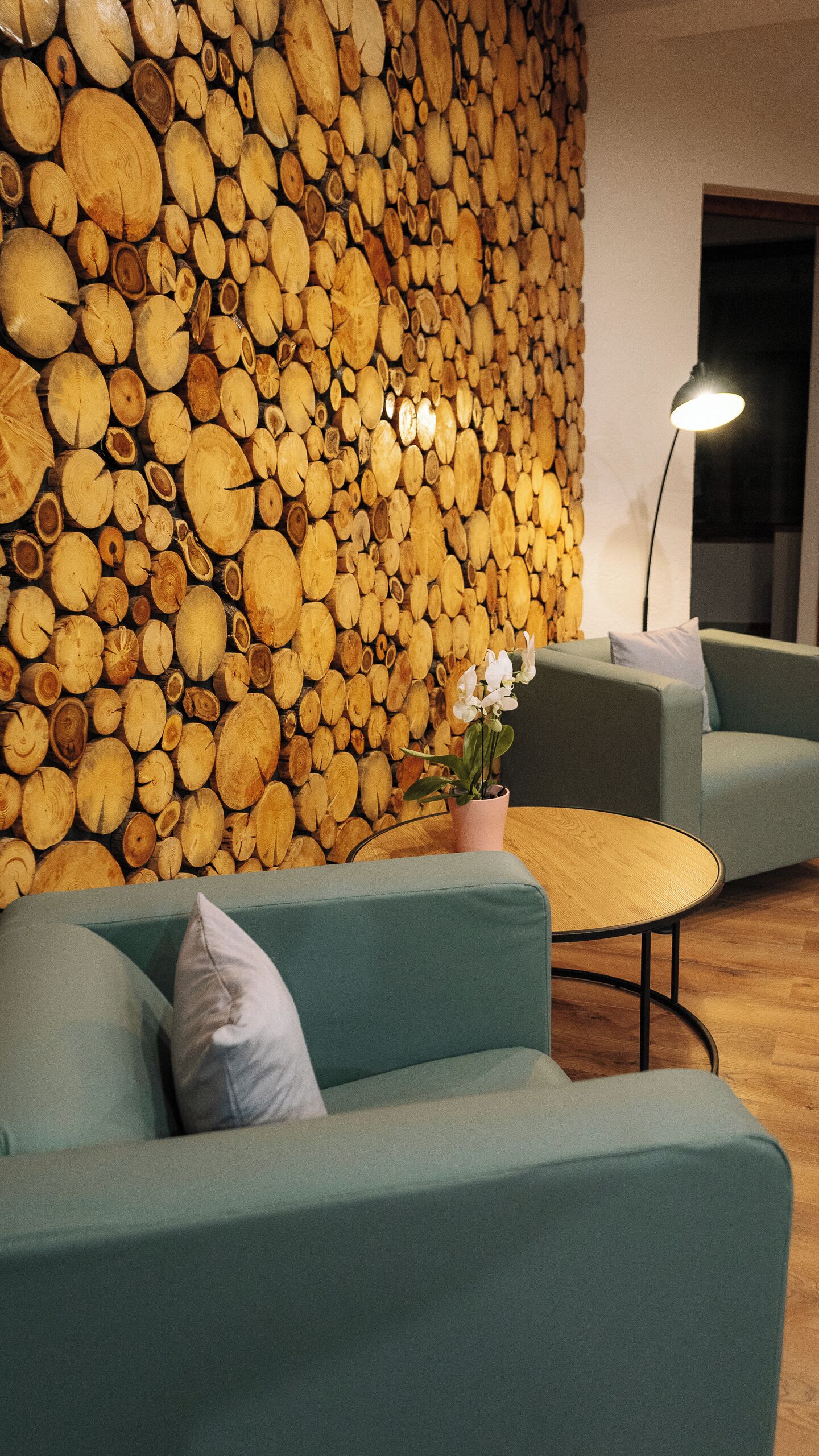 A modern living room with a wall of stacked logs, a green sofa, a small table with a potted plant, and a floor lamp.